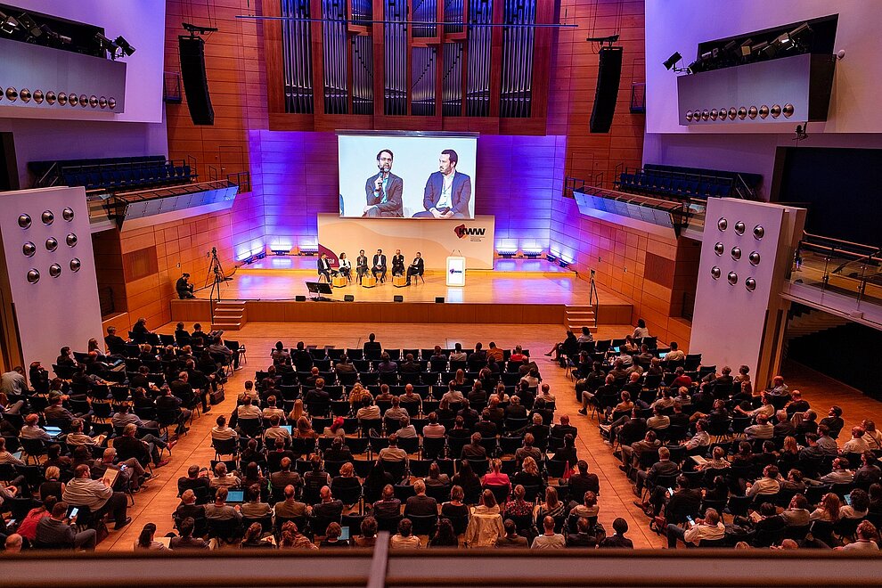 Konferenzsaal mit Publikum und fünf Personen auf der Bühne