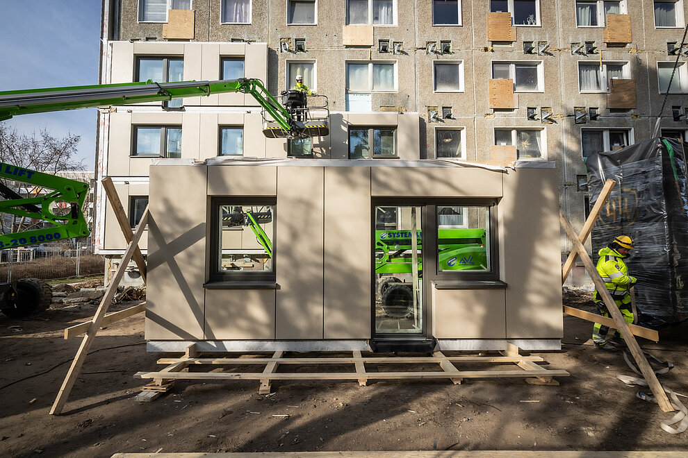 Vorgefertigtes Hausfassadenelement mit Fenstern und Tür. Im Hintergrund Plattenbau während serieller Sanierung