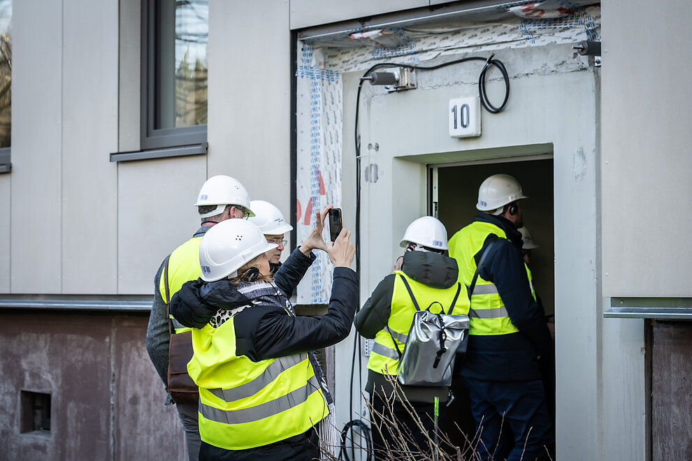 Personengruppe mit gelben Warnwesten und Bauhelmen auf dem Kopf betritt Hauseingang.