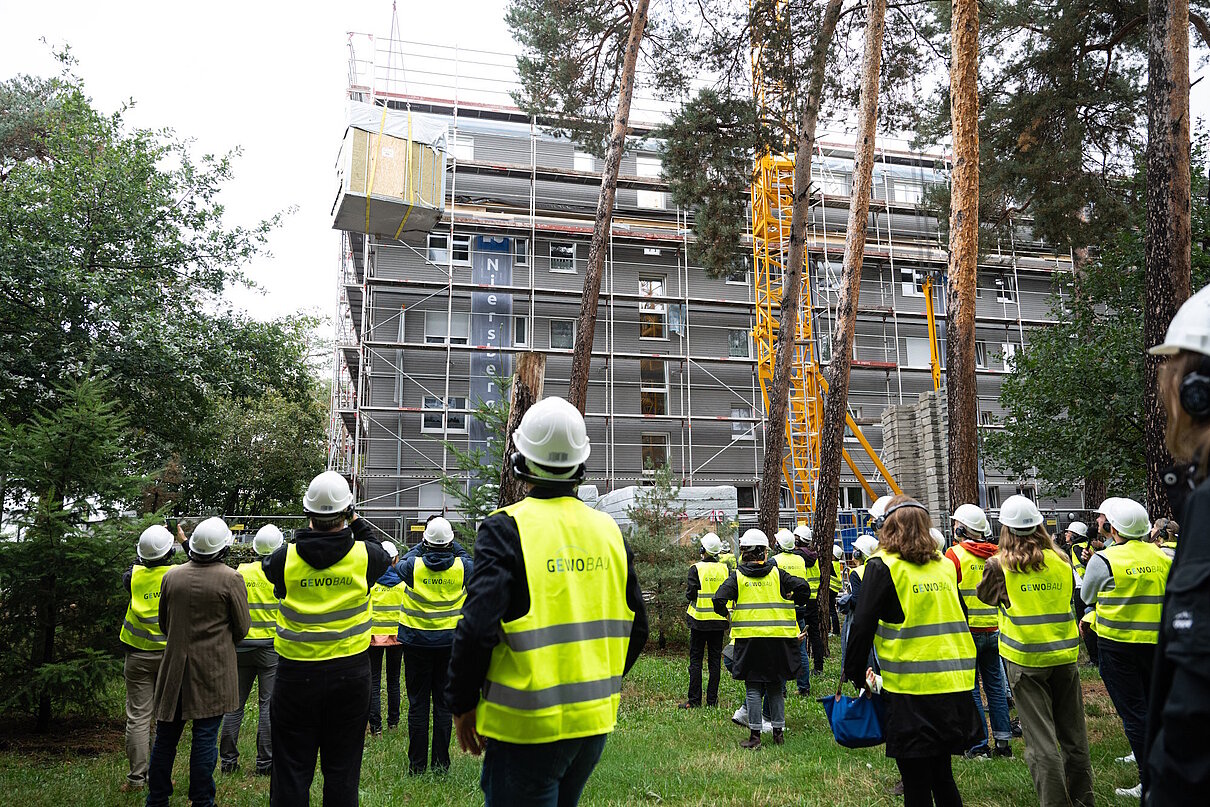 Mehrere Personen mit Warnweste und Helm auf dem Kop auf einer Baustelle blicken zum schwebenden Bauelemente.