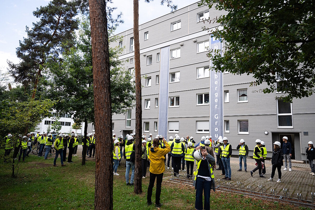 Personen besichtigen eine Baustelle. Im Hintergrund ein Mehrfamliienhaus, davor Rasen und Bäume