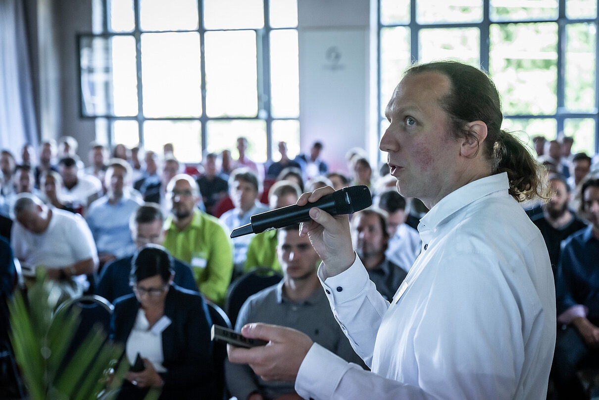 Person mit Mikrofon in der Hand spricht vor Teilnehmenden einer Fachveranstaltung.