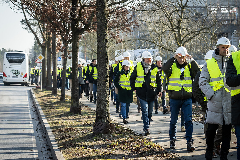 Viele Personen mit gelben Warnwesten und Bauhelmen auf dem Kopf laufen hintereinander auf einem Gehweg.