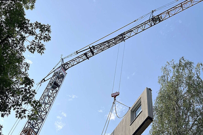 Vorgefertigtes Fassadenelement schwebt an einem Kran über einem Haus, im Hintergrund blauer Himmel.