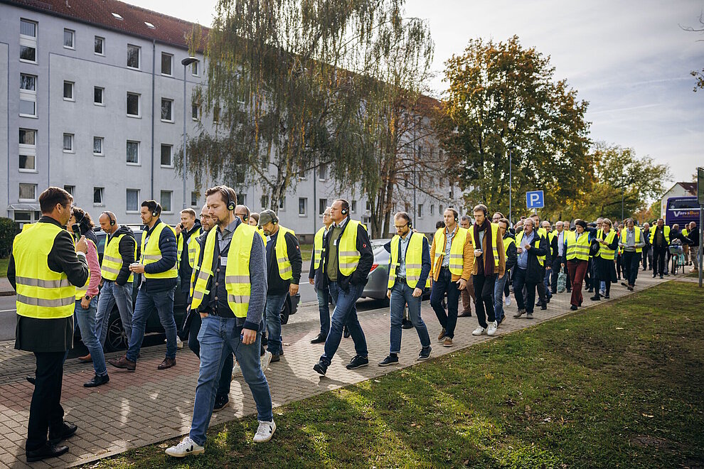 Mehrere Personen in gelben Sicherheitswesten laufen hintereinander auf einem FUßweg, im Hintergrund ist ein Mehrfamilienhaus.