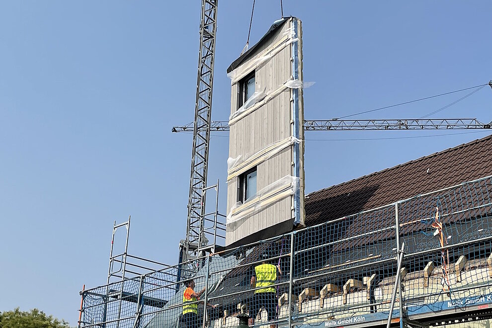 An einem Kran schwebendes Fassadenelement wird von Bauarbeitern zur Montage an Hausfassade platziert.