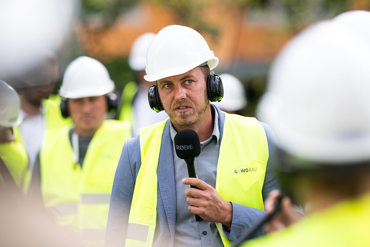 Person mit gelber Warnweste und Helm auf dem Kopf sowie Mikrofon in der Hand