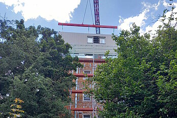 Mehrfamliienhaus mit Baugerüst, schwebendes Fassadenelement an Kran, im Vordergrund Laubbäume und Grünanlagen