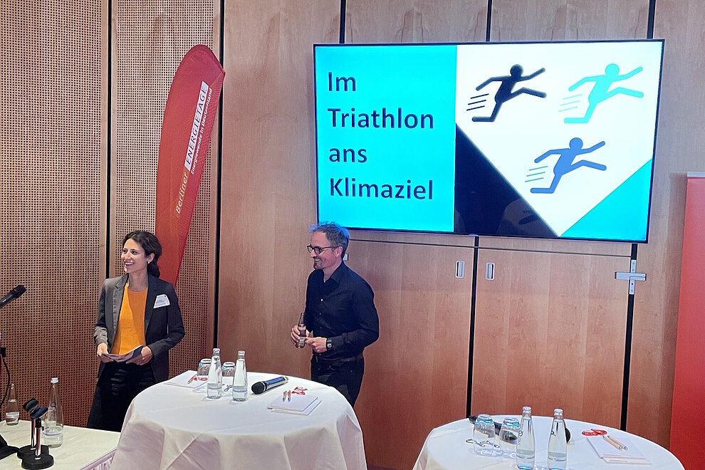Zwei Personen bei einer Fachveranstaltung, im Hintergrund ein großer Bildschirm mit dem Text: Im Triathlon ans Klimaziel
