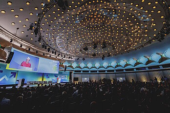 Sitzende Personen in einem Saal bei einem Kongress schauen in Richtung Bühne / Leinwand.