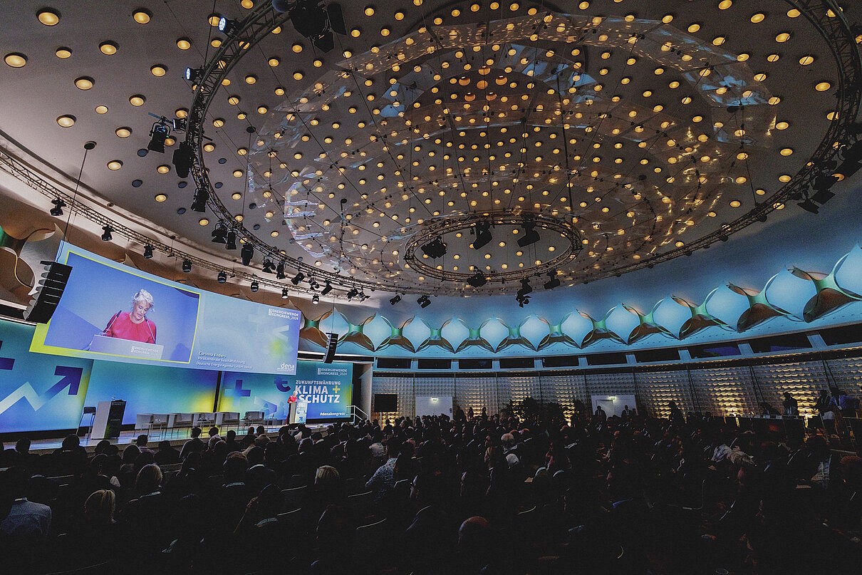 Sitzende Personen in einem Saal bei einem Kongress schauen in Richtung Bühne / Leinwand.