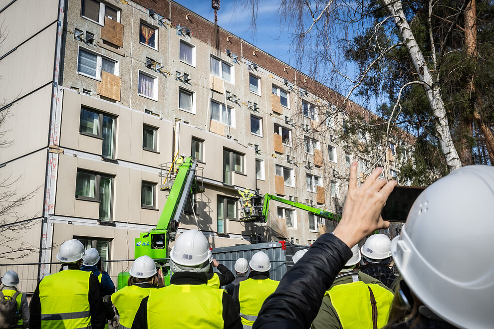 Personengruppe schaut dem Anbringen von Fassadenelementen bei serieller Sanierung eines Plattenbaus zu.