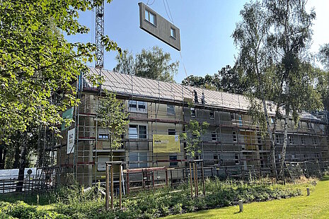 Mehrfamilienhaus mit Schrägdach während Sanierung davor Grünfläche und Laubbäume, Baugerüst, schwebendes vorgefertigtes Fassadenelement zur Montage