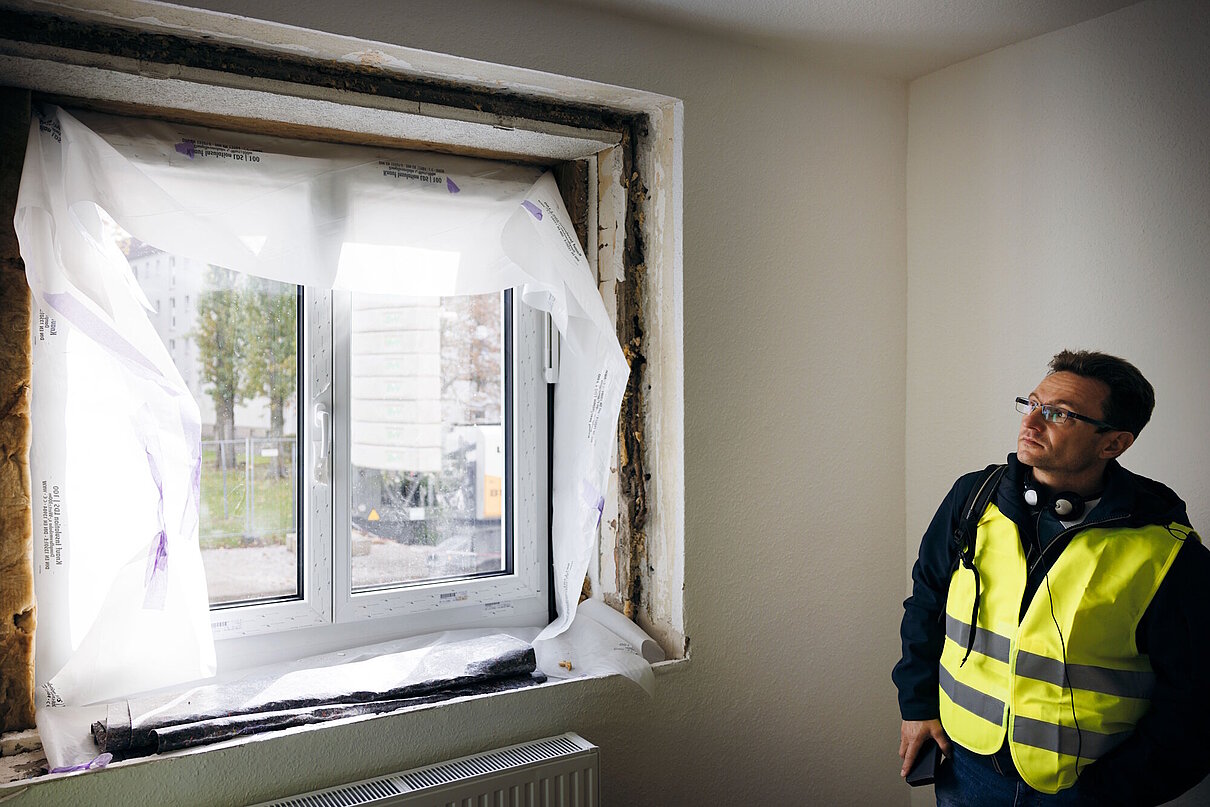 Blick von innen auf ein neu eingesetztes Fenster während einer Sanierung. Seitlich steht eine Person in gelber Sicherheitsweste und betrachtet es.