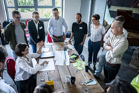 Personengruppe steht um einen großen Holztisch herum, eine Person sitzt, auf dem Tisch liegen Workshop-Unterlagen