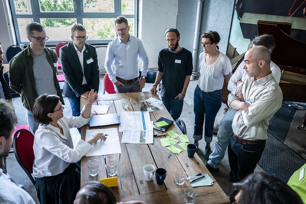 Personengruppe steht um einen großen Holztisch herum, eine Person sitzt, auf dem Tisch liegen Workshop-Unterlagen