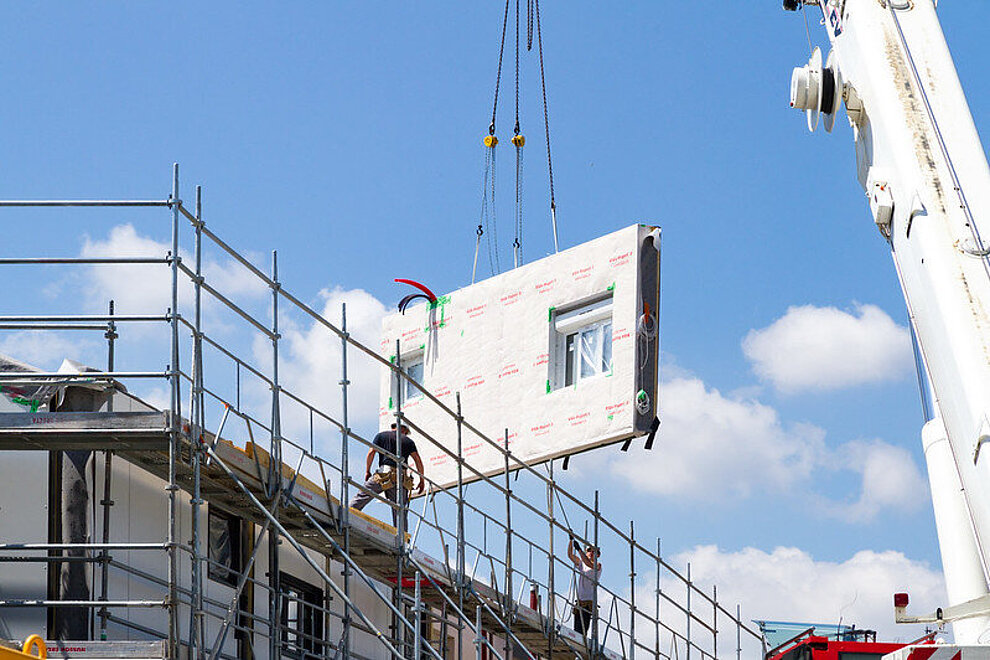 Vorgefertigtes Hausfassadenelement schwebt an einem Kran, Haus mit Baugerüst, zwei Bauarbeiter