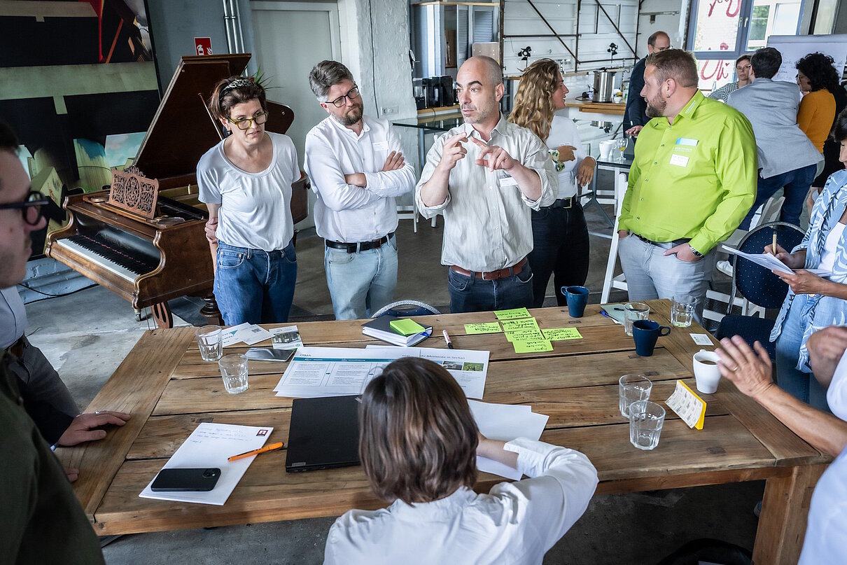 Personengruppe steht während eines Workshops um einen Tisch herum.