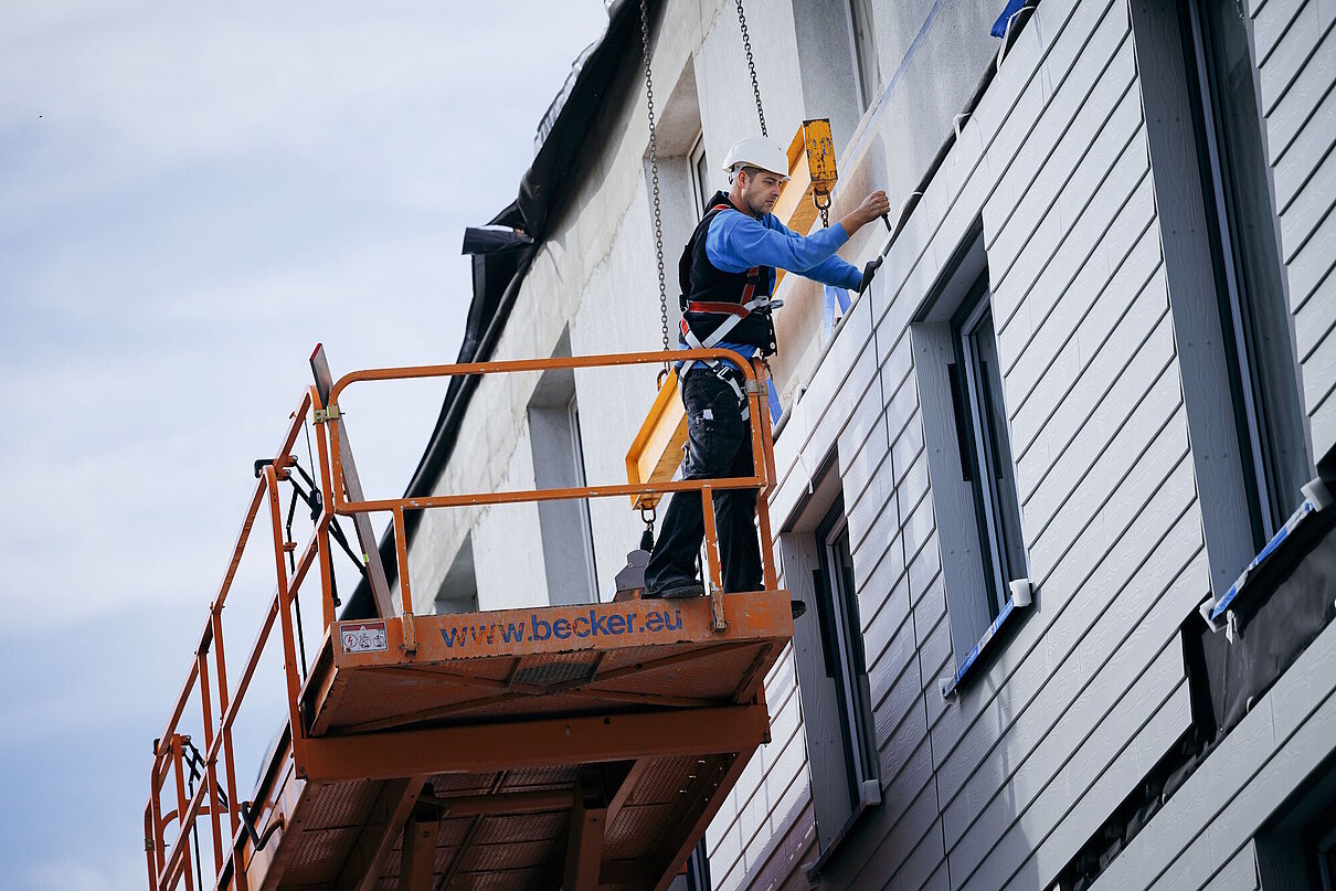 Hausfassade, an die ein vorgefertigtes Fassadenelement durch einen Bauarbeiter montiert wird.